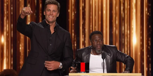 Two men on a stage at an award show: one in a black suit points toward the audience, the other sits at a podium by a microphone, gold curtains in the background.