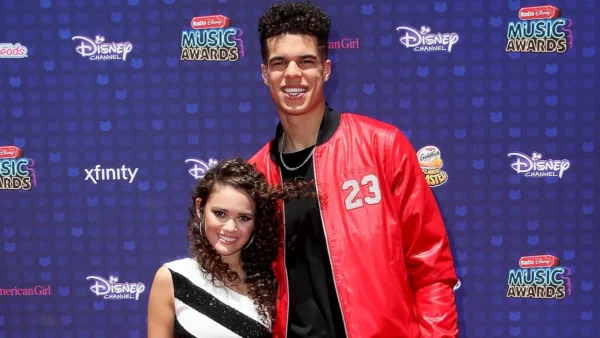Two teens posing on a Disney Music Awards blue carpet; tall guy in a red jacket with the number 23 and a chain, a girl in a black-and-white striped dress next to him.