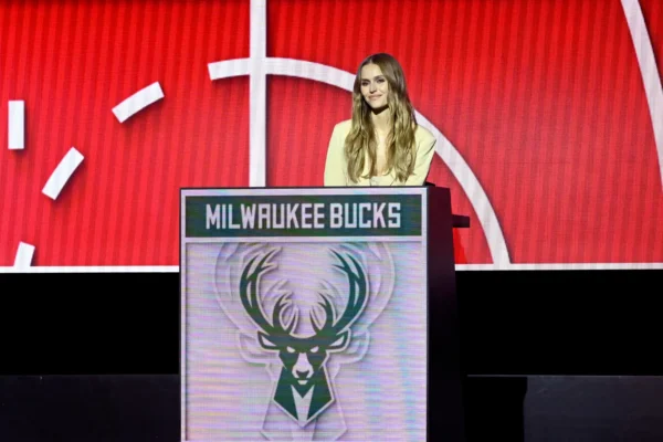 Woman with long blond hair in a light blazer stands at a podium with a Milwaukee Bucks logo on the front, red stage backdrop behind her.