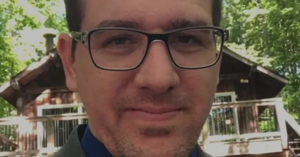 Man with dark hair and glasses close-up outdoors, a cabin in the background.