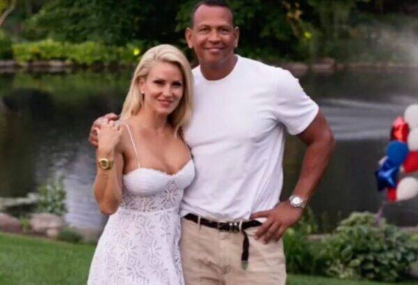 Smiling couple posing outdoors by a pond; woman in a white lace dress, man in a white t-shirt and khaki shorts.
