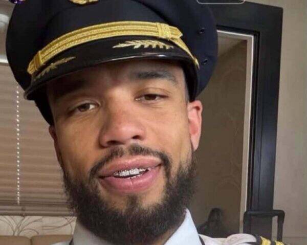 Close-up of a man with a beard and braces wearing a navy captain's hat with gold braid, smiling at the camera indoors.