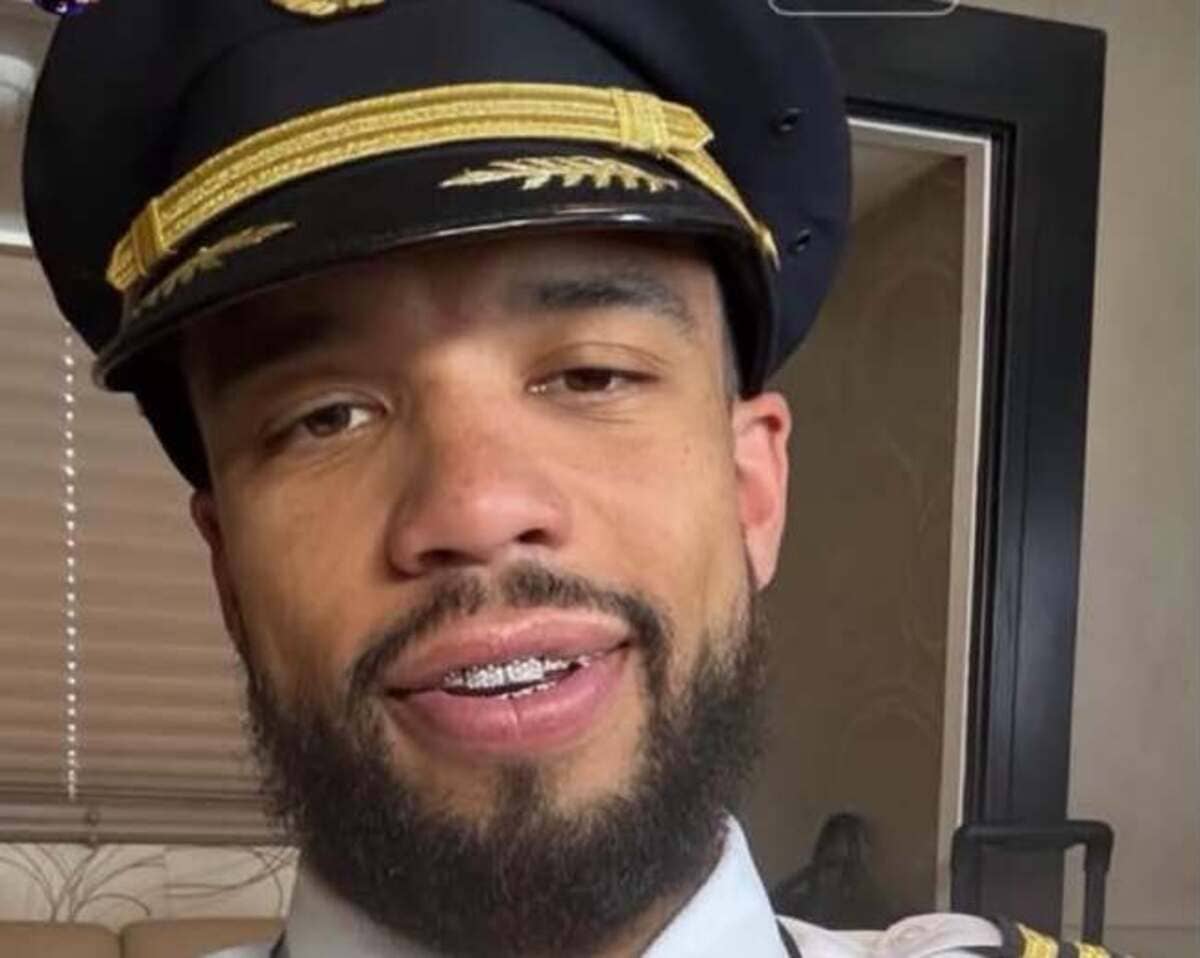 Close-up of a man with a beard and braces wearing a navy captain's hat with gold braid, smiling at the camera indoors.