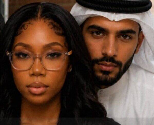 Close-up of a woman wearing clear-framed glasses with a man in a white kandura and headscarf behind her.
