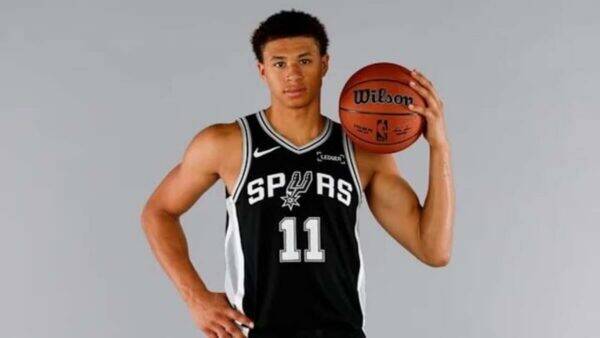 Male basketball player in a Spurs jersey (#11) holding a basketball against a gray studio background.