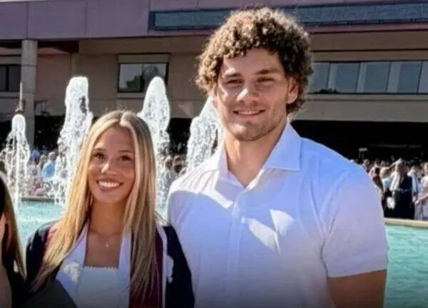 Two friends posing by a fountain; the woman wears a graduation gown and white top, smiling at the camera.