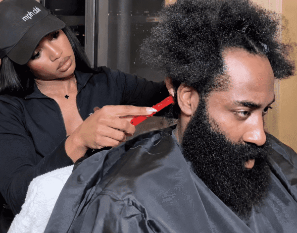 Stylist trimming a man's afro with a red comb while he wears a black cape in a salon.