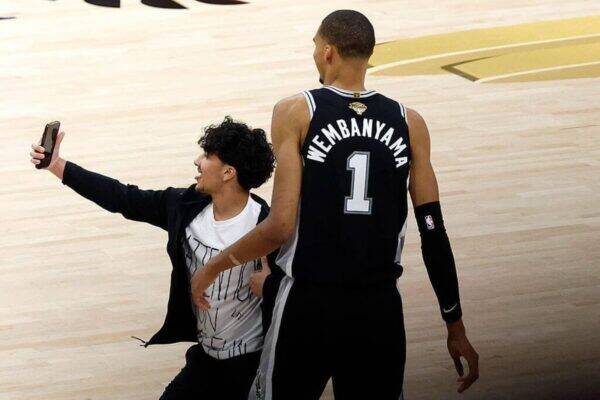 Two basketball players on a light wooden court; one in a Wembanyama #1 jersey stands while a friend in a jacket takes a selfie with a smartphone.