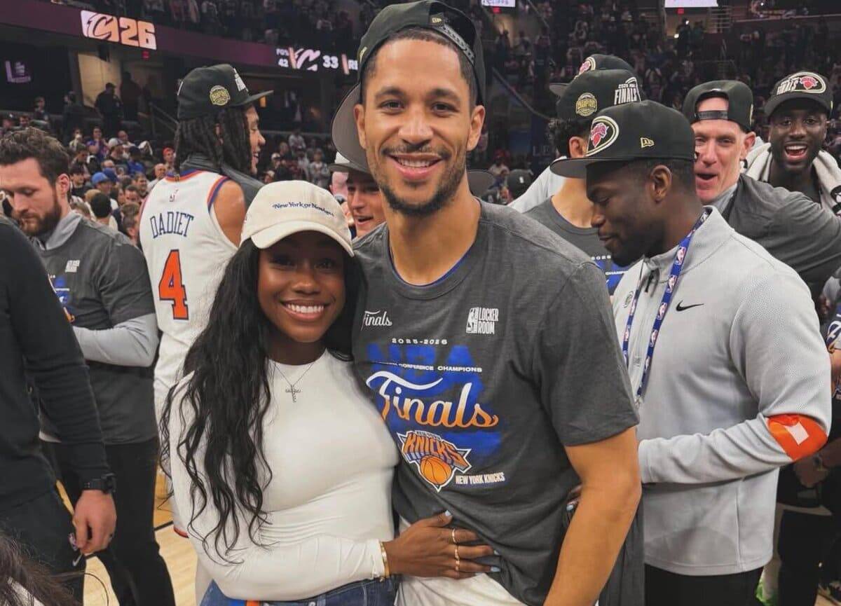 Smiling couple hugs on a basketball court, man in a Knicks Finals shirt and woman in white with a cap, teammates celebrate around them.