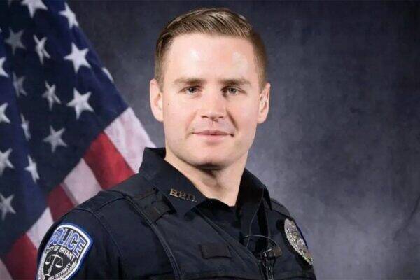 Portrait of a male police officer in uniform with badge, standing in front of an American flag.