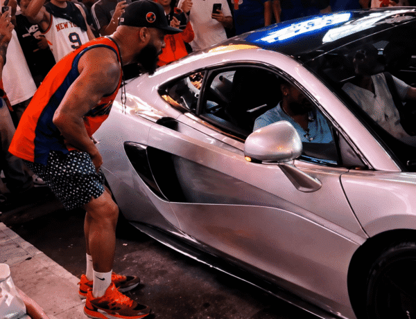 Man in an orange jersey and patterned shorts leans toward a silver sports car as a crowd watches and photographs in the background.