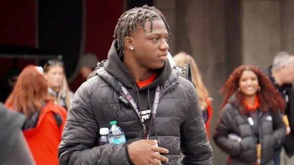 Young man with braided hair wearing a black puffer jacket and lanyard, holding a water bottle, looking to the side at an outdoor event.