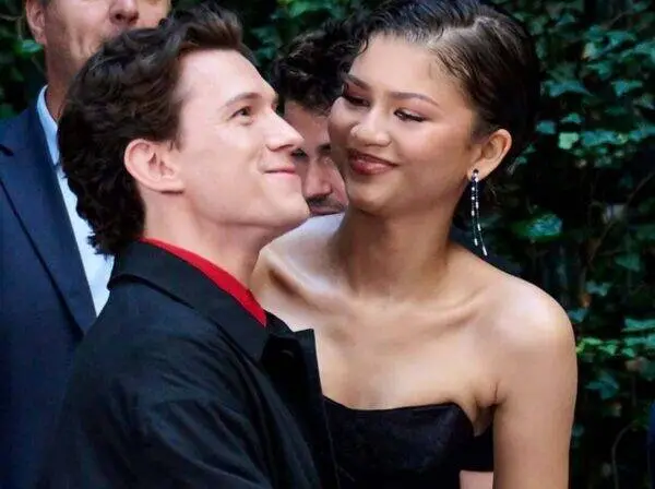 Couple in formal attire smiling at each other outdoors, greenery behind them.