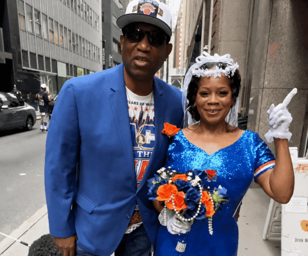 Couple posing on a city street; man in a blue blazer and cap, woman in a sparkly blue dress with a veil holding a bouquet and raising a gloved finger.