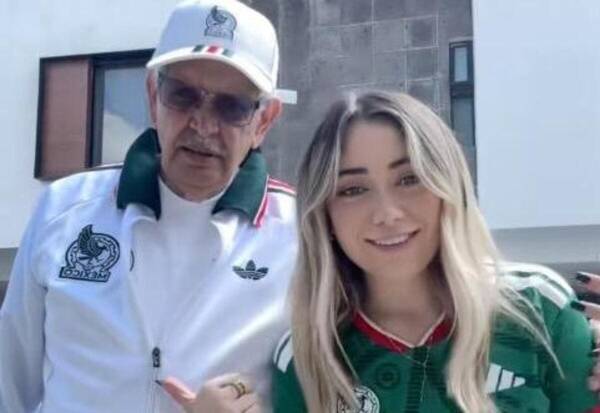Two people posing outdoors and smiling: a man in a white jacket with green collar and a cap, beside a smiling woman in a green jersey with blonde hair on the right.