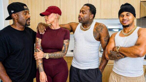 Four friends stand in a kitchen with arms around each other, dressed in casual athletic wear, chatting warmly.