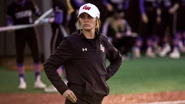 Female athlete wearing a GW white cap and black jacket stands with hands on hips on a softball/baseball field, looking to the side.