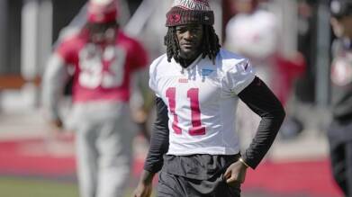 Football player in white 49ers jersey number 11 walking on the field wearing a red beanie and black sleeves.