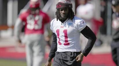 Football player in white 49ers jersey number 11 walking on the field wearing a red beanie and black sleeves.