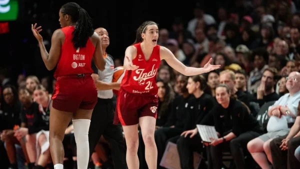 Two Indiana Fever players in red uniforms on a basketball court; one gestures with arms wide as the other faces away while the crowd watches the game.