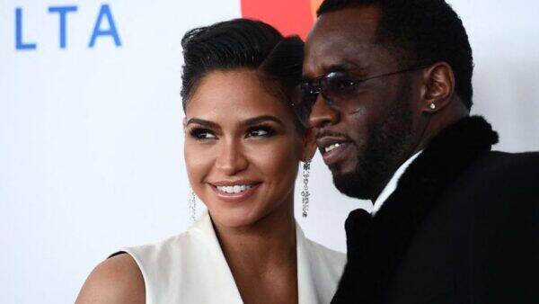 Smiling woman in a white sleeveless dress with chandelier earrings posing with a man in a black suit at a red carpet event before a light backdrop.
