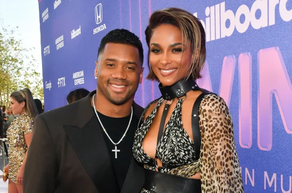 Smiling couple posing together on a purple Billboard backdrop at a red-carpet event; man in a black suit with a pearl necklace, woman in a leopard-print outfit with a black choker and cutout top.