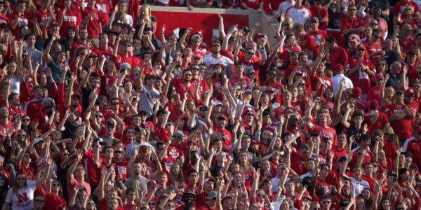 Large crowd of sports fans in red, raising hands and cheering at a stadium in celebration.