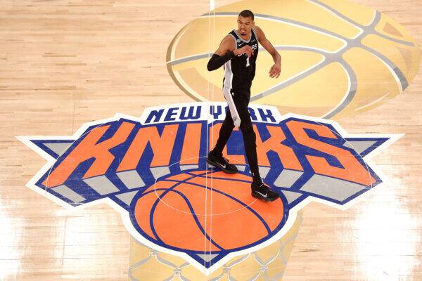 Basketball player in a black Spurs uniform jumps mid-air on a hardwood court over a large New York Knicks logo.