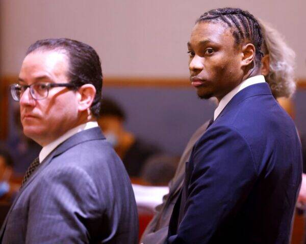 Two men in formal suits in a courtroom; one with glasses facing left, the other glancing toward the camera.