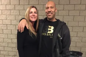A smiling man and woman stand side by side against a cinder block wall, the man's arm around the woman's shoulders.
