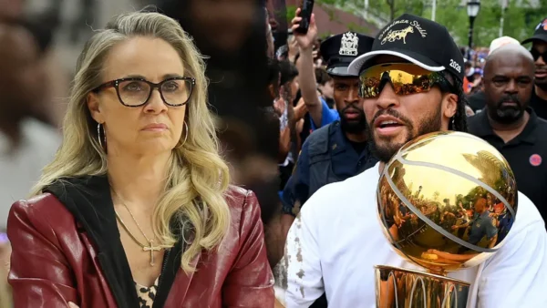 Woman with blonde hair and glasses sits with a serious expression beside a man wearing mirrored sunglasses, holding a large gold basketball trophy with a crowd behind them.