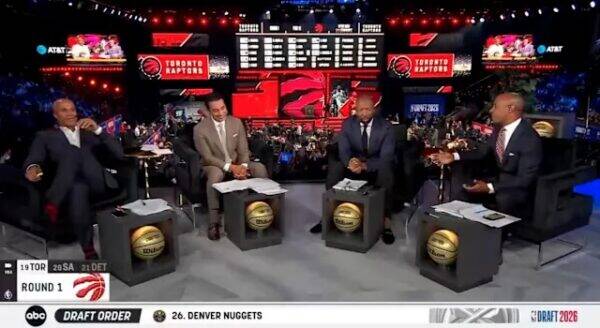 Four male commentators sit at podium desks on a basketball draft show set, with Raptors branding and a crowded arena backdrop.