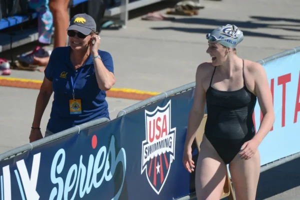 Swimmer in a black one-piece and swim cap smiles as she walks beside a blue barrier banner labeled USA Swimming.