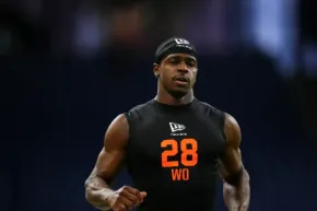 Male athlete in a black sleeveless shirt with bright orange 28 and WO text, wearing a black headband, mid-run on a dark background.