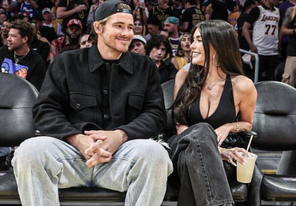Couple sitting courtside at a sports event, smiling at each other; man in a black jacket and cap, woman in a black top holding a drink.