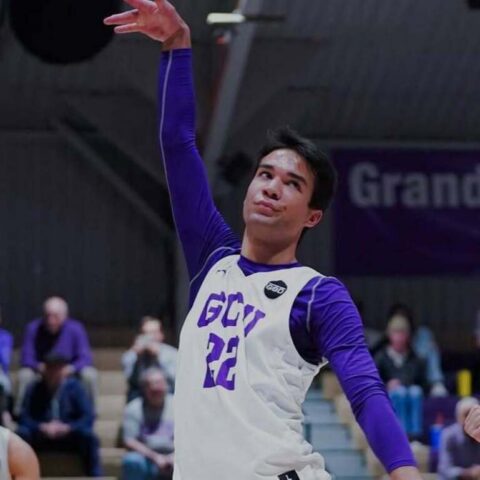 Basketball player in a white jersey with purple sleeves (#22) leaps with arm raised during a game, crowd in the background.