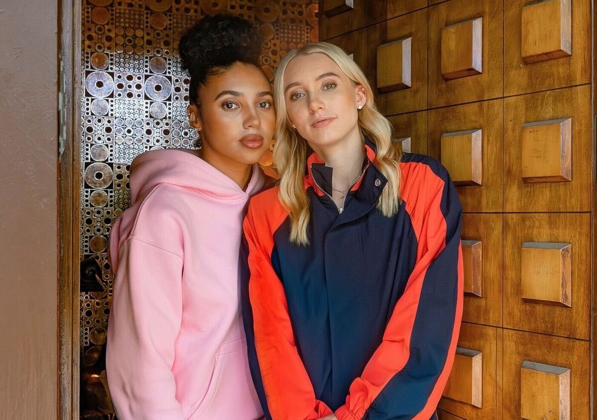 Two women stand close together indoors: one in a pink hoodie, the other in a navy and red windbreaker, both looking at the camera.