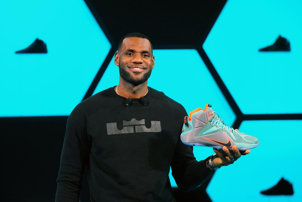 Man on a stage holding a teal sneaker with orange accents, against a bright blue geometric backdrop.