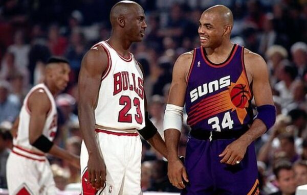 Michael Jordan in a Bulls uniform and Charles Barkley in a Suns uniform smile at each other on the court.