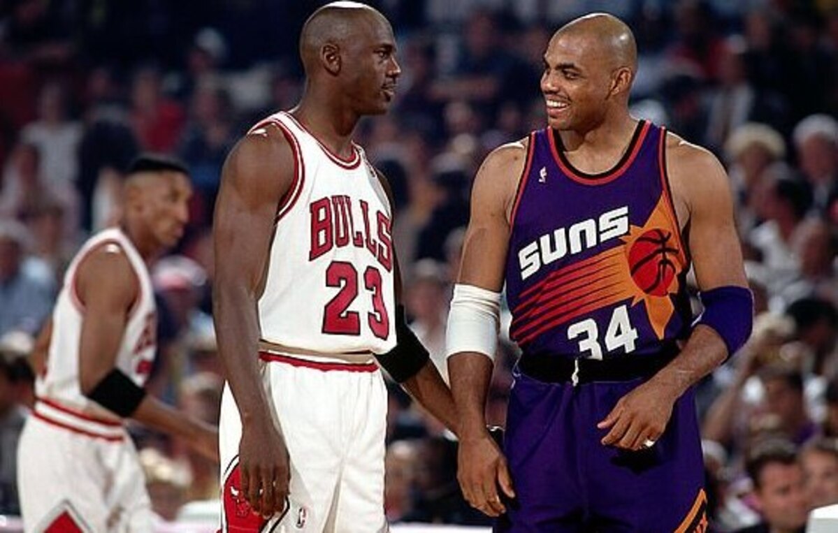 Michael Jordan in a Bulls uniform and Charles Barkley in a Suns uniform smile at each other on the court.