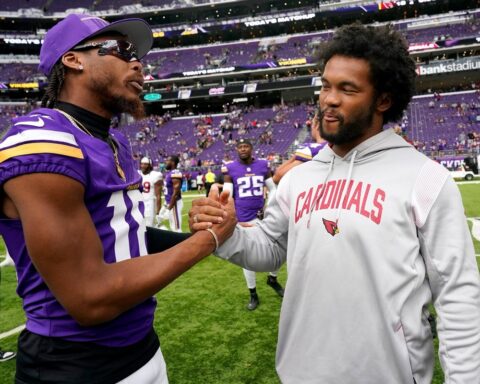 Two football players shake hands on the field, one in a purple Vikings uniform and the other in a gray Cardinals hoodie, with purple stadium seats behind them.