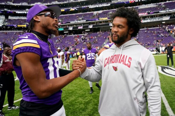 Two football players shake hands on the field, one in a purple Vikings uniform and the other in a gray Cardinals hoodie, with purple stadium seats behind them.
