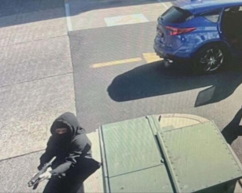 Masked armed individual in a black hoodie holding a handgun on a sidewalk near a blue SUV with its door open, beside a green dumpster.