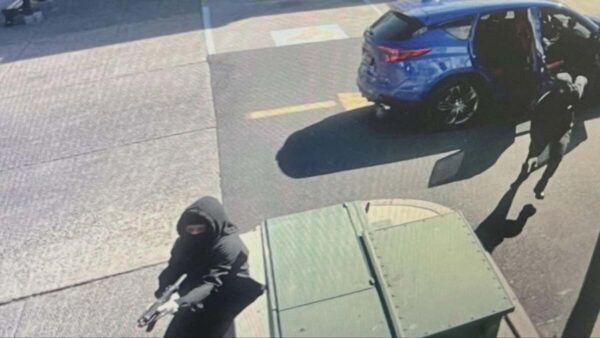 Masked armed individual in a black hoodie holding a handgun on a sidewalk near a blue SUV with its door open, beside a green dumpster.