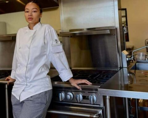 Female chef in a white jacket leaning on a stainless steel commercial stove in a professional kitchen.