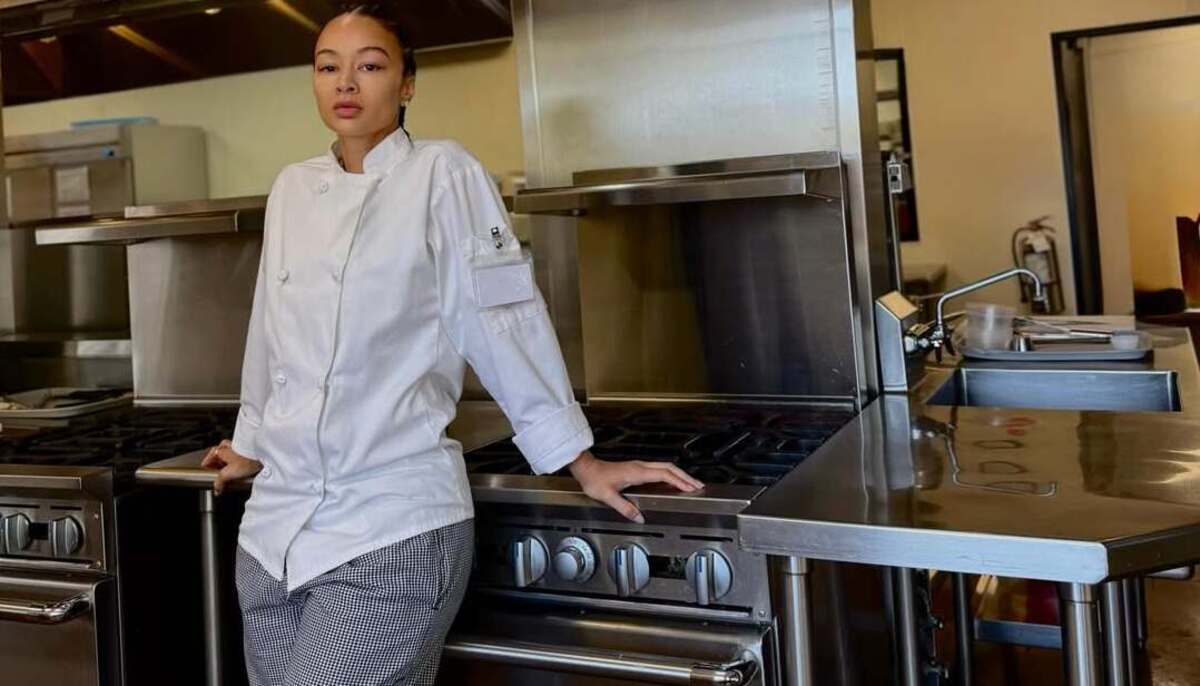 Female chef in a white jacket leaning on a stainless steel commercial stove in a professional kitchen.
