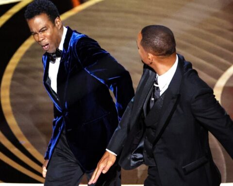 Two men in formal tuxedos dance on a stage with a circular gold backdrop behind them.