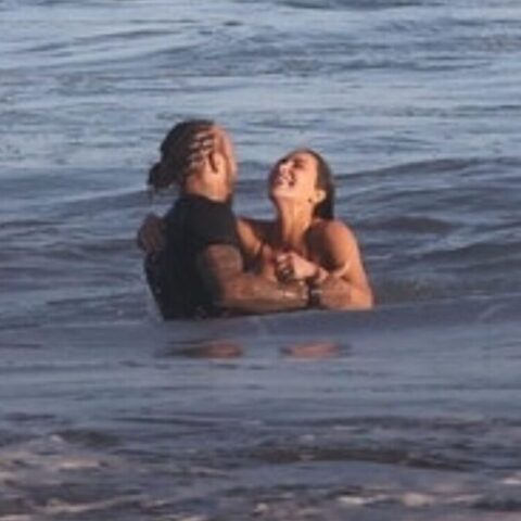 Two people standing waist-deep in the ocean embrace each other in the surf, facing and leaning toward one another.
