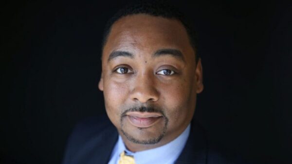 Close-up headshot of a man in a navy suit and yellow tie, looking at the camera against a dark background.