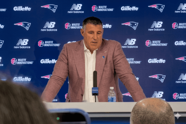 Man in a pink blazer speaks at a press conference, blue sponsor backdrop behind him; Deere-labeled mic on the podium.